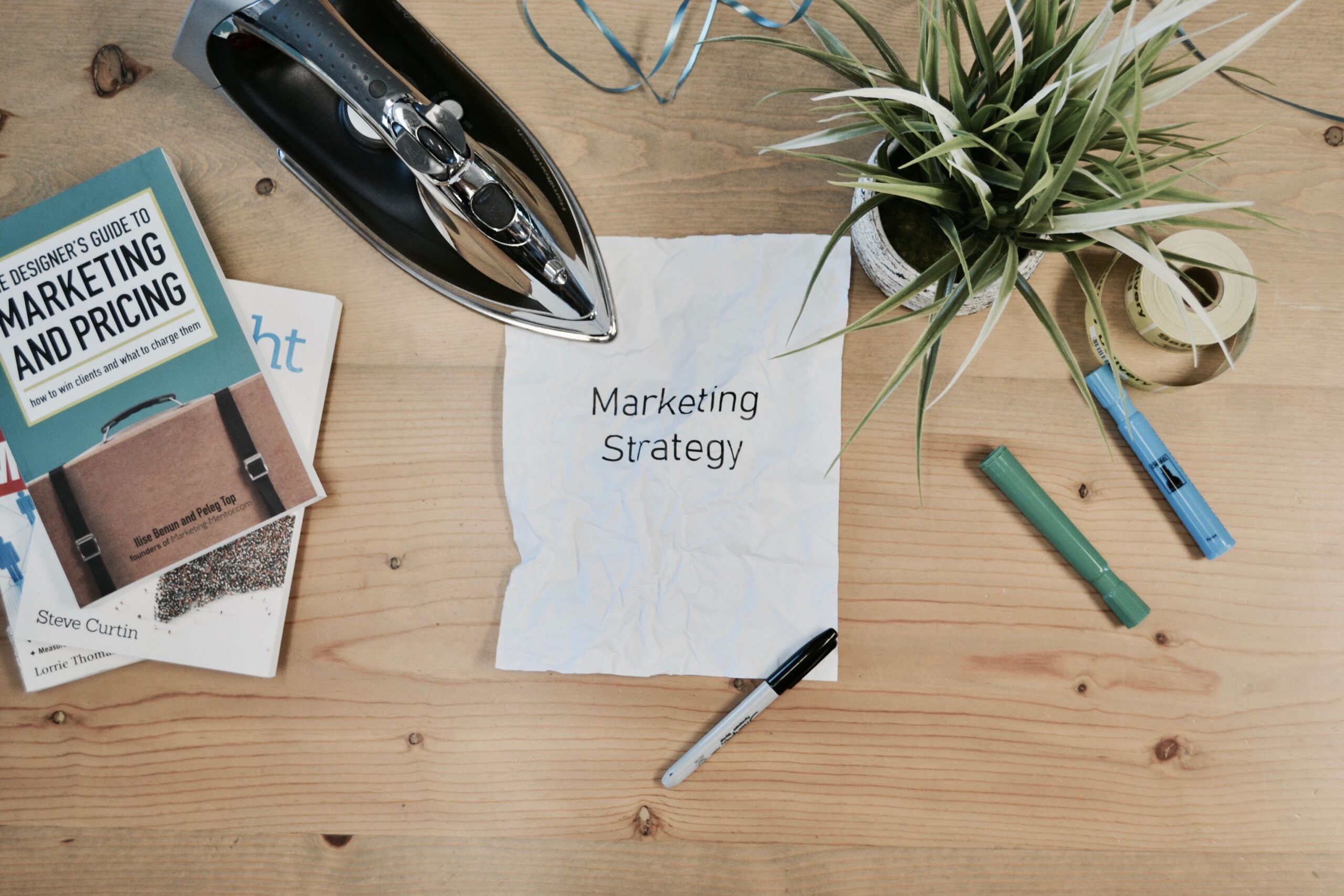 Holztisch mit Schreibutensilien, einer Pflanze und einem Bügeleisen, das auf einem zerknitterten Papier mit dem Schriftzug „Marketing Strategy“ steht