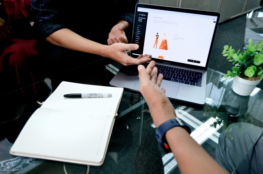 Aufgeklappter Laptop auf einem Glastisch, zwei Personen sitzen darum und zeigen auf den PC